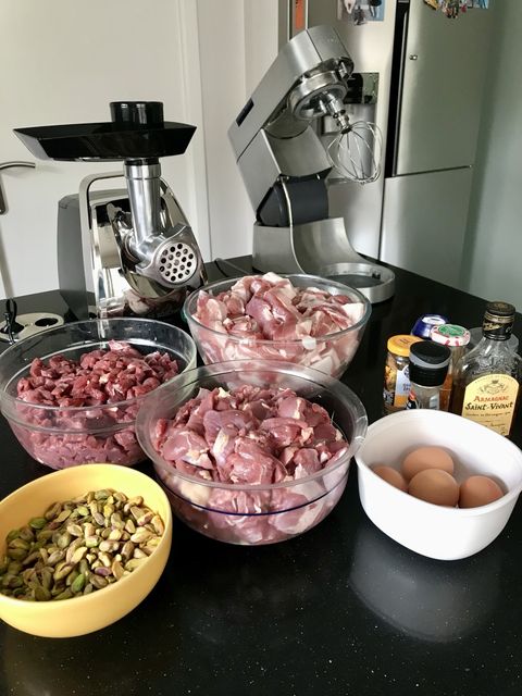 Mise en place for a pâté: bowls of cubed pork and game, a bowl of pistachios, eggs, spices and a bottle of armagnac, with a meat grinder and stand mixer behind